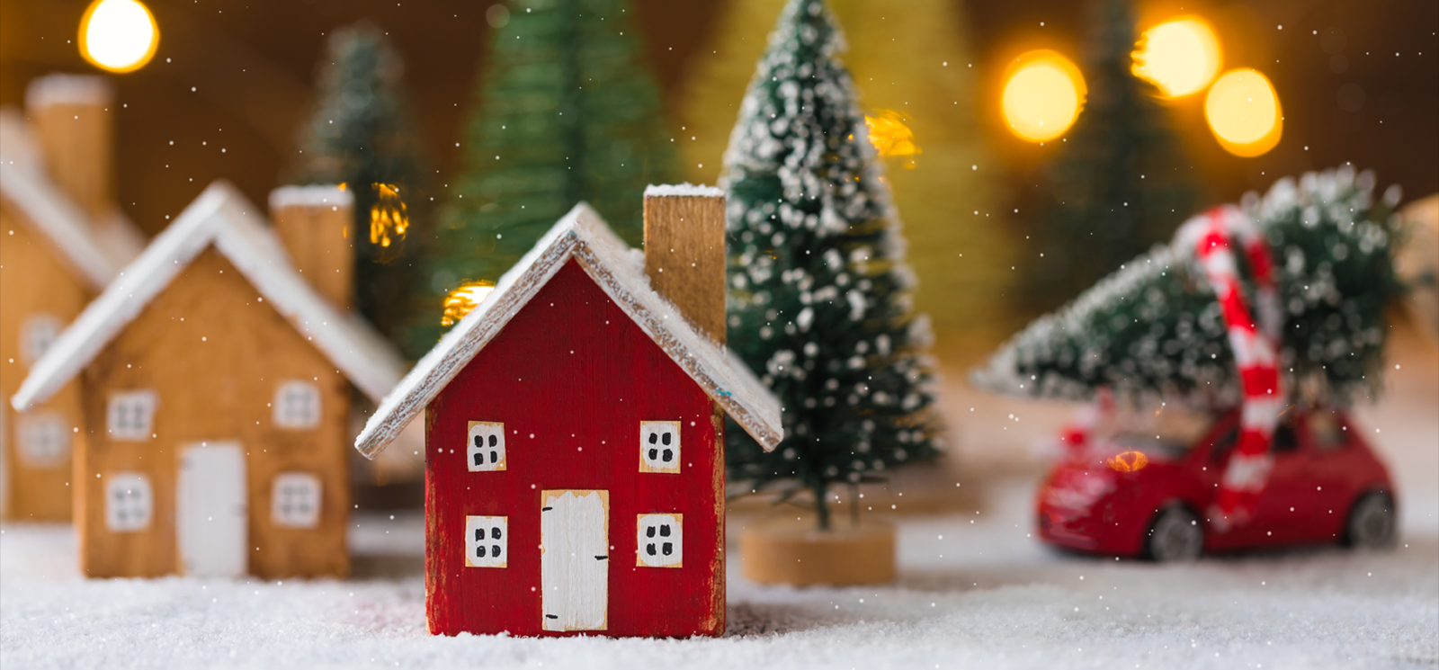 Miniature wooden houses and small red car with fir tree on the snow over blurred Christmas decoration background